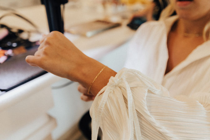 Close-up of a person in a white tie-sleeve blouse wearing a delicate gold bracelet while using a black hair-styling tool at a vanity.