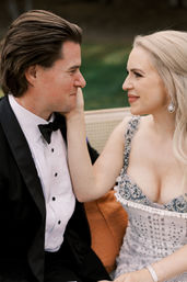 Man in black tuxedo and woman in beaded gown smile at each other as she gently cups his cheek while seated on an outdoor garden bench