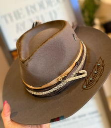 Hand holding a brown felt western-style fedora with a wide brim, tan and rhinestone hatband secured by a jeweled safety pin and leather strap, engraved feather motif with turquoise studs on the brim.