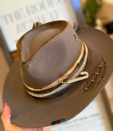 Hand holding a brown felt western-style fedora with a wide brim, tan and rhinestone hatband secured by a jeweled safety pin and leather strap, engraved feather motif with turquoise studs on the brim.