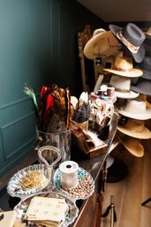 Close-up of a hat shop display: glass jars of colorful feathers, trays with beads, chains and a spool of trim on a counter, and a rack of wide-brimmed hats in the background.