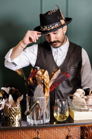 Stylish man in a leather vest tipping a feathered fedora behind a cocktail bar, jars of decorative feathers and a glass of amber drink on the counter