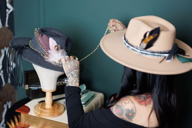Tattooed artisan wearing a beige fedora adjusts a feather-and-chain decorated gray felt hat on a mannequin head against a teal wall.