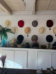 Sunlit boutique hat shop interior with colorful fedoras and straw cowboy hats mounted on a white wall, stacked on a counter under rustic wood beams and accented by a turquoise vase with a fern