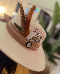 Close-up of a beige felt fedora with a tan ribbon, layered brown feathers and a king and queen playing card tucked into the hatband — playful boho hat accent.