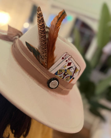 Close-up of a beige felt fedora with a tan ribbon, layered brown feathers and a king and queen playing card tucked into the hatband — playful boho hat accent.