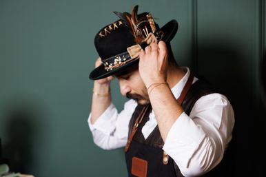Man in white shirt and leather apron adjusts a stylish black felt hat decorated with feathers and stitched trim against a green wall