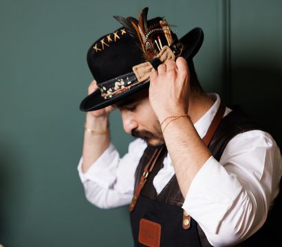 Man in white shirt and leather apron adjusts a stylish black felt hat decorated with feathers and stitched trim against a green wall