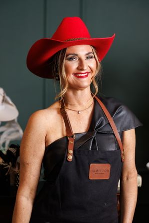 Smiling woman wearing a vibrant red cowboy hat, leather one-shoulder top and leather-strap apron in a hat boutique studio