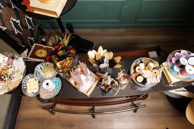 Overhead view of a craft workstation on a curved wooden console table — a playful mix of colorful feathers, beads, buttons, wooden spools and rolls of ribbon in bowls and trays.