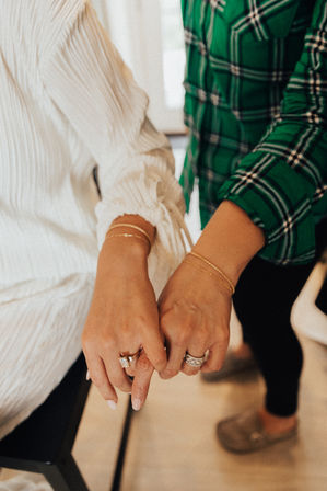 Close-up of two people linking pinky fingers, showing diamond rings and delicate gold bracelets; one wearing a white blouse and the other a green plaid shirt in a cozy indoor setting.