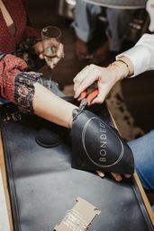 Close-up in a jewelry boutique: artisan uses red-handled pliers to cut a delicate gold bracelet from a woman's wrist over a black leather mat while she holds a champagne flute.