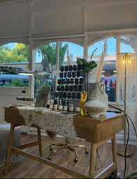 Sunlit boho-chic boutique desk with wooden table displaying small cosmetic jars and black product cards on a tiered rack, gold hand sculpture, ceramic vases, potted plant, and beaded floor lamp by arched windows.