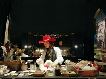 Person wearing a bright red cowboy hat arranging hats and accessories on a boutique counter filled with ribbons, feathers, boxes and jewelry under warm shop lighting