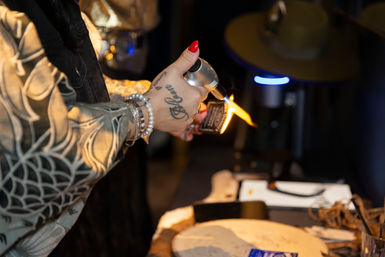 Close-up of a tattooed hand with red nails using a culinary torch to flame a small item over a wooden serving board in a dim restaurant or bar setting.