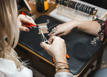 Close-up of hands using red-handled pliers to repair a silver chain necklace on a jewelry workbench, with bracelets and tools nearby