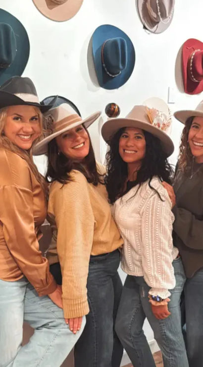 Four smiling friends trying on wide-brim felt hats in a boutique, posing in front of a white wall with colorful hat displays while wearing casual fall outfits and denim.