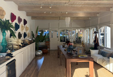 Cozy sunlit boutique hat shop interior with colorful hats mounted on a white wall, wooden-beamed ceiling, central wooden display table with accessories, cushioned sofa seating and large arched windows.