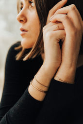 Close-up portrait of a woman in a black top wearing delicate gold ring and layered gold bracelets, hands gently clasped near her face in soft natural light.