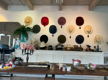 Bright boutique interior with colorful wide-brim and cowboy hats arranged on a white wall under exposed wooden beams, counter stacked with hat forms, and a front table displaying ribbons, trims, and craft supplies.