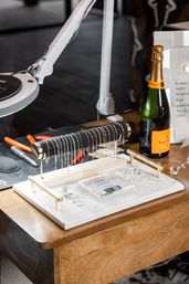 Small jewelry workspace with a marble tray holding a black T-bar displaying delicate silver chains, scattered crystal accents, pliers and lamp on a wooden table next to a champagne bottle.