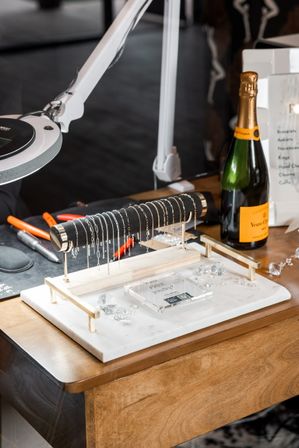 Small jewelry workspace with a marble tray holding a black T-bar displaying delicate silver chains, scattered crystal accents, pliers and lamp on a wooden table next to a champagne bottle.