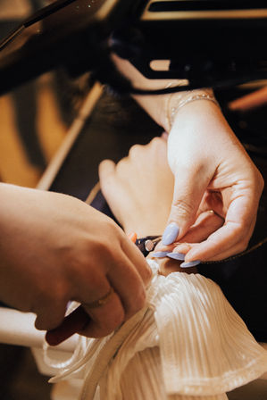 Close-up of manicured hands with lavender nails fastening a clasp on a pleated white wedding dress during a bridal alteration or fitting