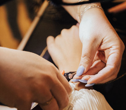 Close-up of manicured hands with lavender nails fastening a clasp on a pleated white wedding dress during a bridal alteration or fitting