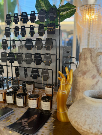 Artisan market booth table with rows of dangling earrings on black display cards, amber glass candles, a gold hand ring holder wearing rings, and rustic ceramic vases.