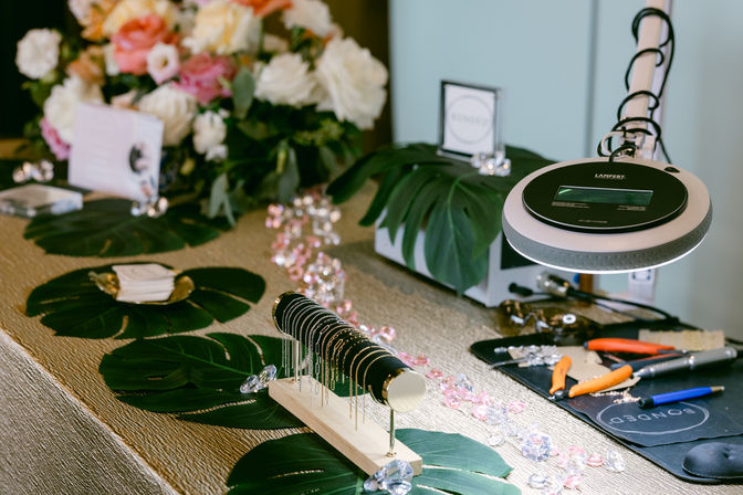 Artisan jewelry display on a textured table: delicate gold and silver bracelets on a black velvet roll, scattered pink crystal gems and large monstera leaves, a magnifying lamp and pliers on a small workbench, with a soft floral arrangement in the background.