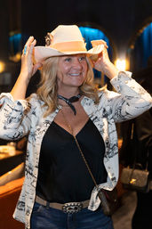 Smiling woman tries on a white cowboy hat in a dimly lit lounge, wearing a printed jacket, black top, jeans, silver-buckled belt and crossbody bag.