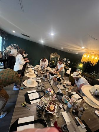 Busy hat-making workshop in a cozy indoor studio — long table strewn with straw wide-brim hats, ribbons, beads, trims and tools as a small group decorates and styles hats under warm chandelier light and a decorative mirror on dark green walls.