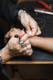 Close-up of a tattooed nail technician shaping a client's nails at a nail salon, hands adorned with rings and bracelets and a manicure tool in use