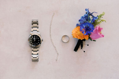 Flat lay of groom’s accessories on pale pink background: stainless steel wristwatch, silver chain and wedding band beside a colorful boutonniere with blue, orange and pink flowers.