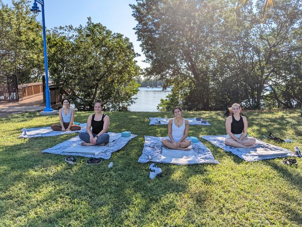 Four people seated cross-legged on patterned yoga mats in a sunny waterfront park lawn, trees framing a calm harbor with sailboats in the background.