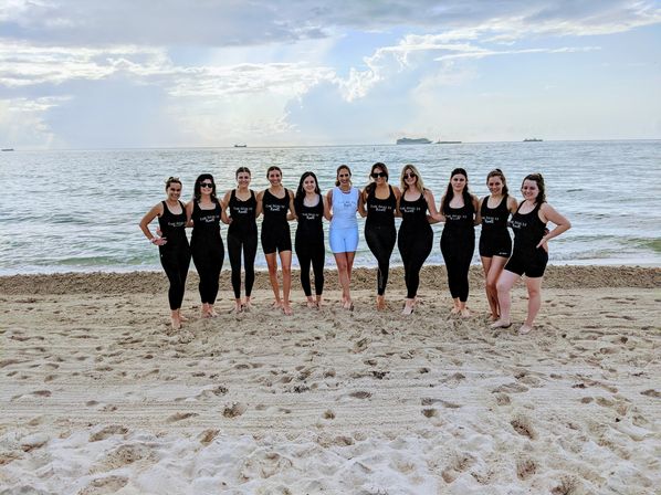 Smiling group of women in matching black swimsuits with one in white, arms linked on a sandy oceanfront beach with calm sea, ships on the horizon and a dramatic cloudy sky — seaside group photo, beach and coastal scenery.