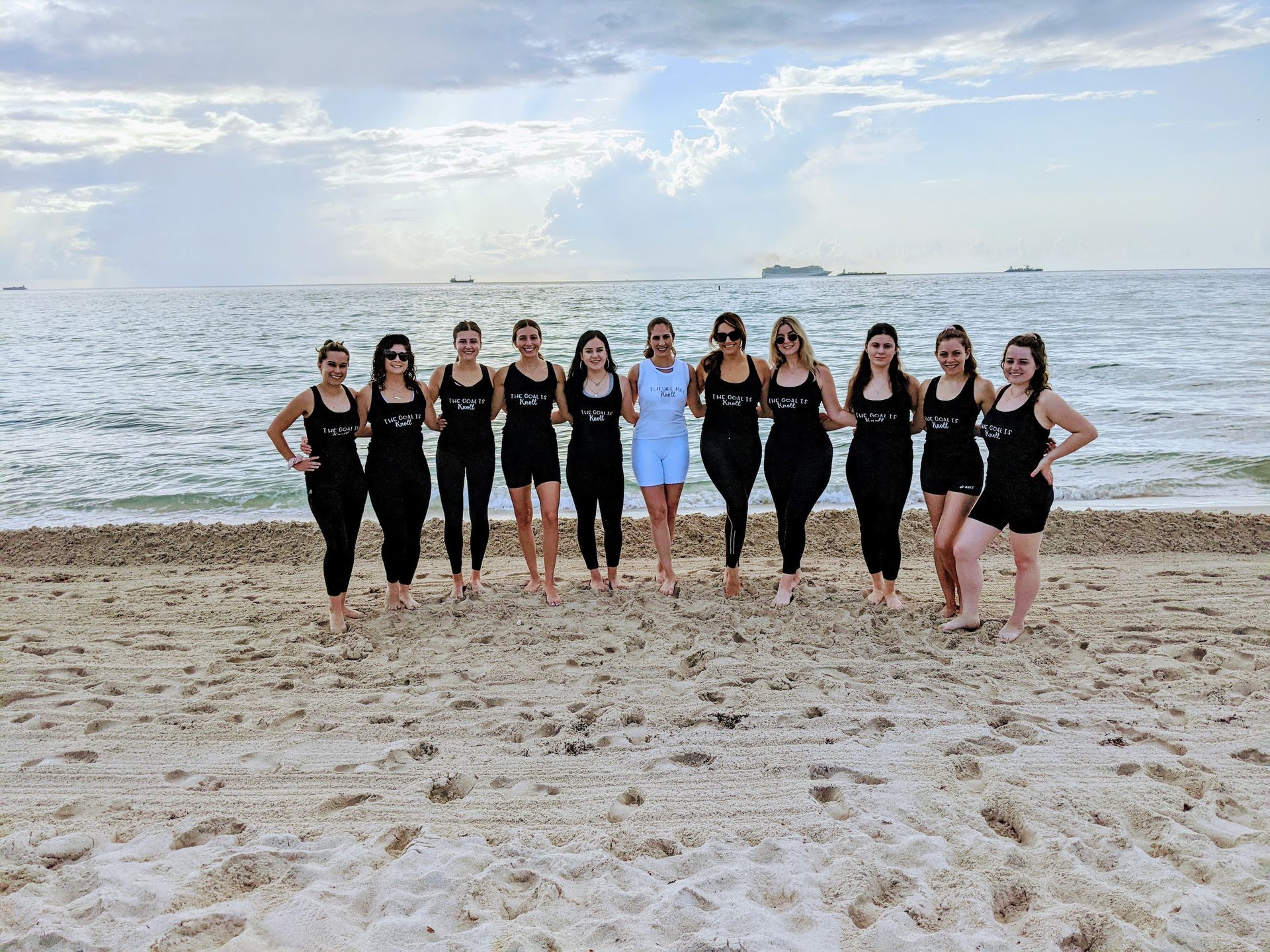 Group of women in matching black swimsuits and one in white standing barefoot on a sandy oceanfront beach with linked arms, calm water, ships on the horizon and a sunlit cloudy sky.