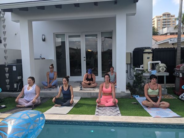 Serene poolside yoga session with several women seated on mats on artificial grass in a suburban backyard beside a modern white house and swimming pool.