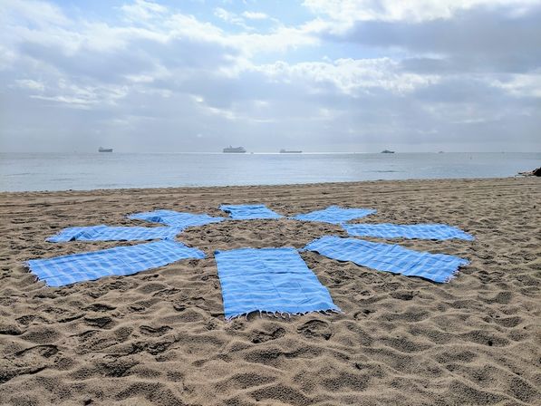 Blue towels arranged in a sunburst on a sandy beach facing a calm sea with cargo ships on the horizon under a cloudy sky