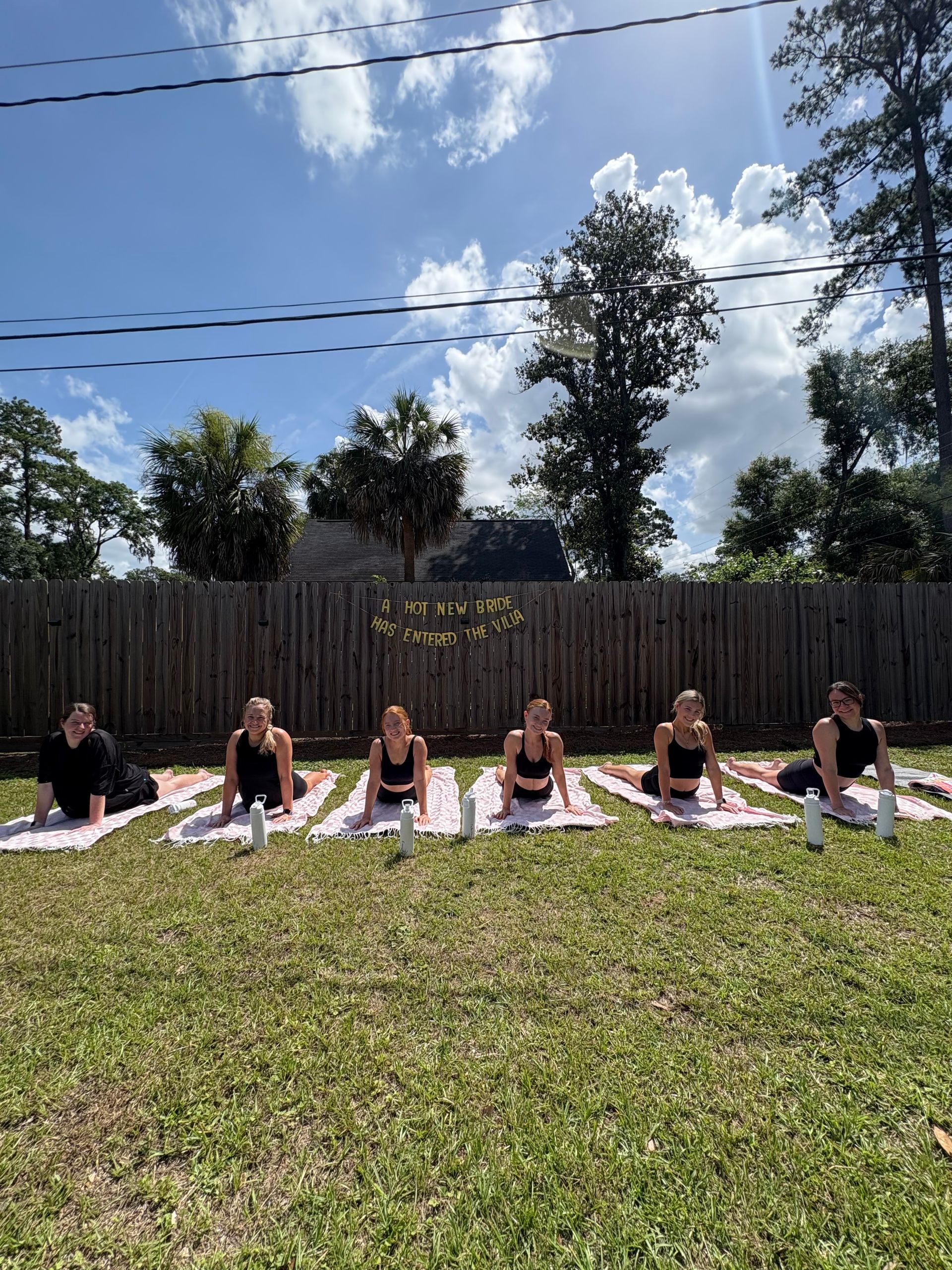 Six women in black activewear doing cobra pose on striped towels for a sunny backyard bachelorette yoga session, with water bottles, palm trees, wooden fence and a celebratory banner in the background.