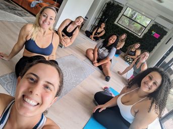 Smiling group of women in activewear sitting on yoga mats in a bright, plant-lined studio, posing for a post-class selfie after a group fitness/yoga session.
