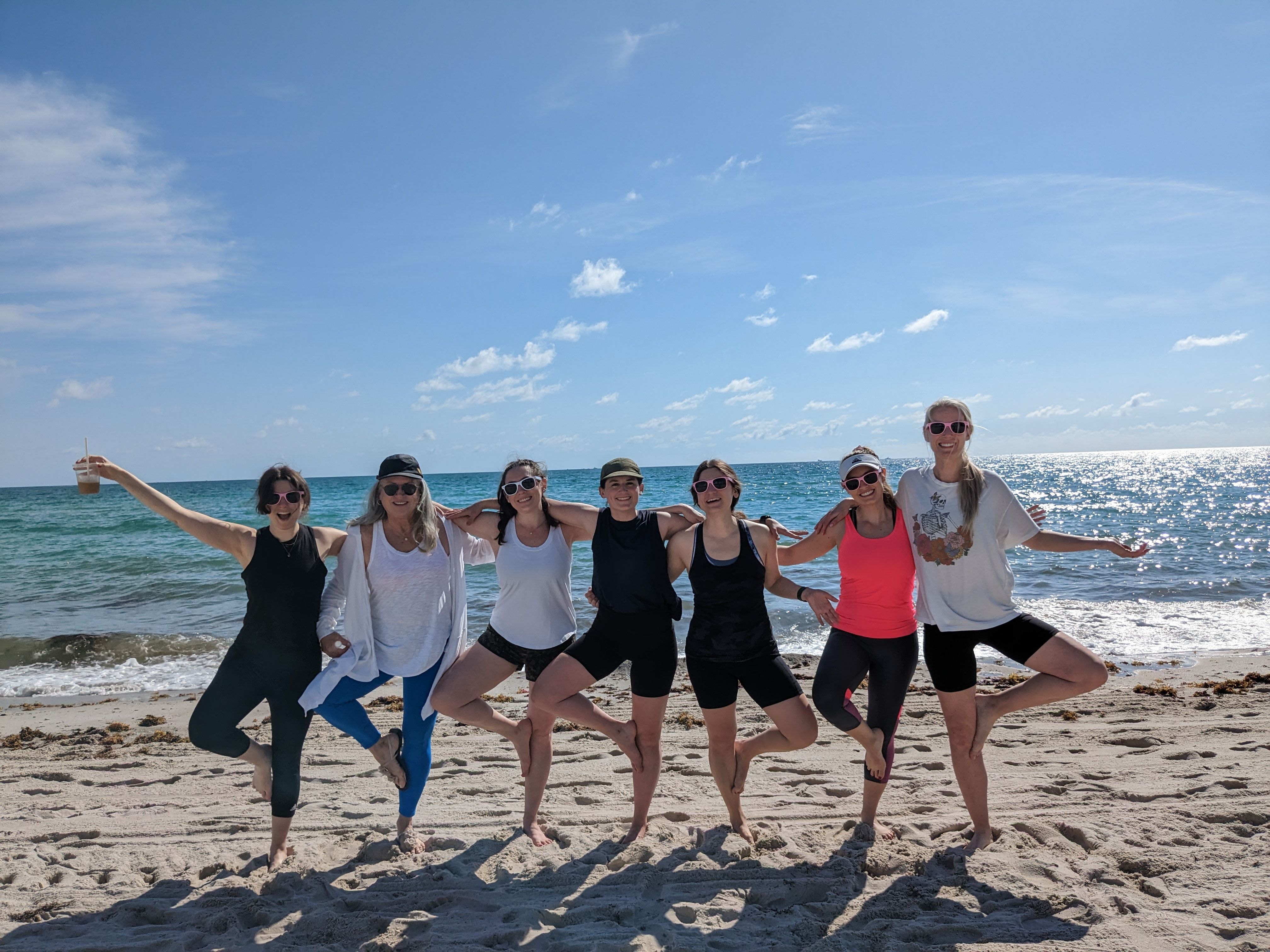 Seven women in athletic wear and sunglasses pose arm‑in‑arm in tree pose on a sunny sandy beach with sparkling blue ocean and clear sky.