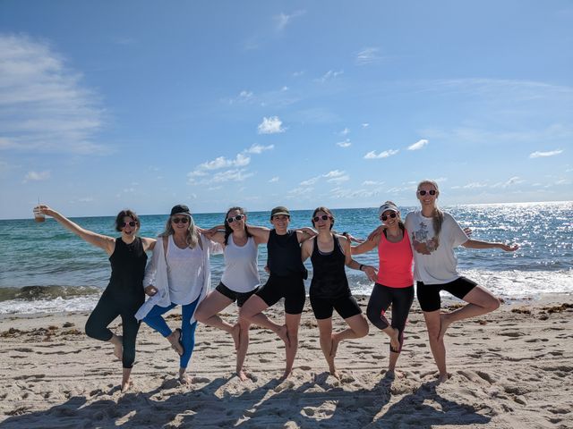Group of eight people doing tree-pose yoga on a sunny sandy beach, arms linked with turquoise ocean and clear blue sky in the background, most wearing sunglasses and activewear.