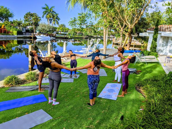 Seven people holding hands in a circle practicing outdoor yoga on mats on a sunny, palm-lined residential waterfront lawn beside a calm canal with docks and a boat.