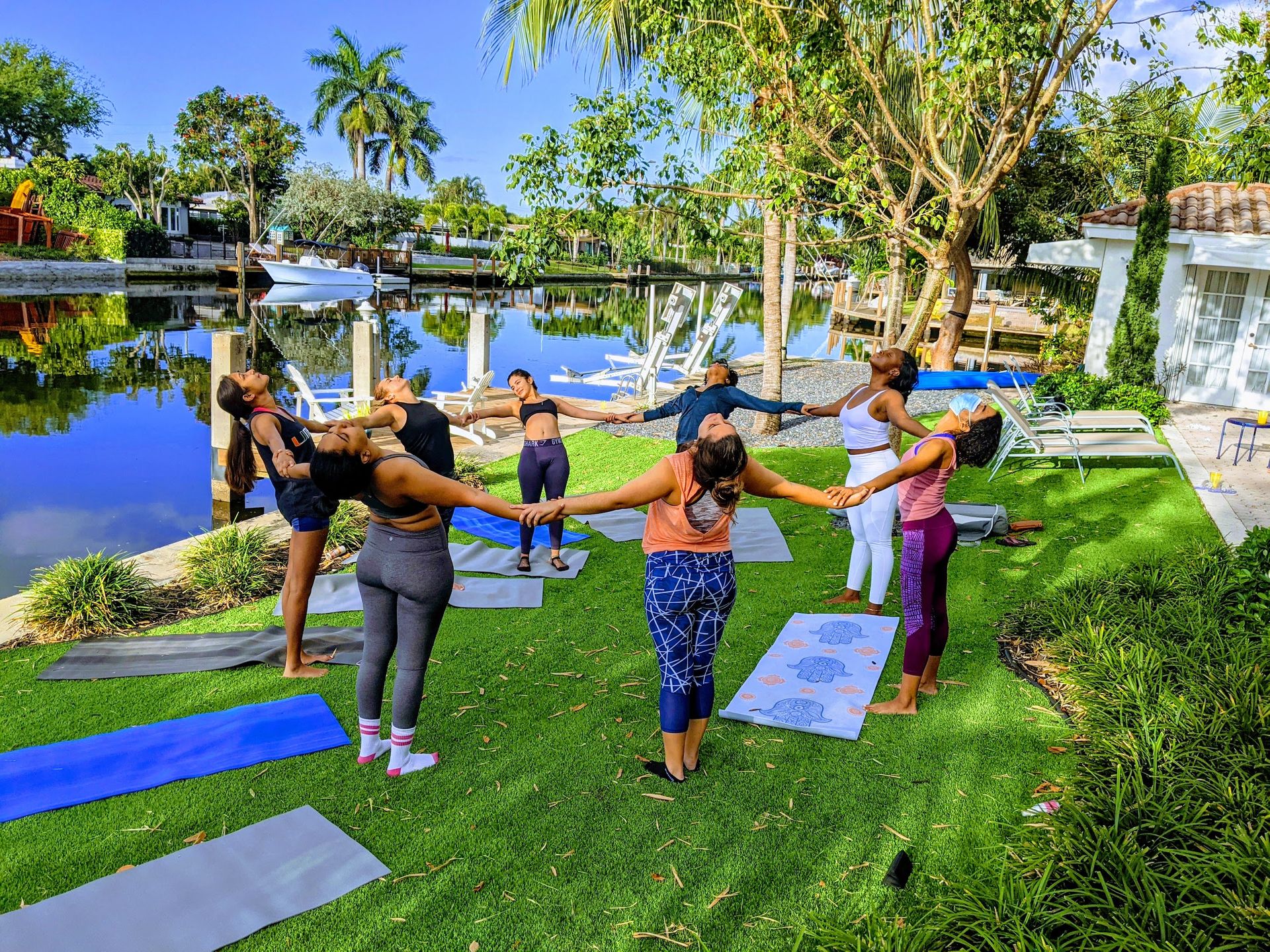 Outdoor group yoga circle on a sunny tropical waterfront canal lawn; women holding hands on mats by palm trees and docks.