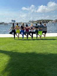 Eight people in colorful activewear doing tree-pose with arms around each other on a grassy lawn facing a sunny waterfront marina with boats and palm trees.