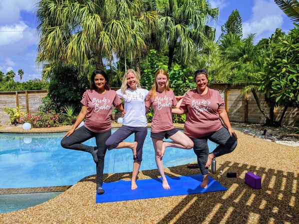 Four women in matching 'Bride's Babes' shirts practicing tree pose on a blue yoga mat beside a sunny backyard pool surrounded by palm trees.