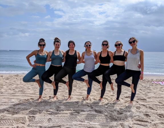 Seven women in activewear linked arm-in-arm practicing tree pose for a group beach yoga session on sandy shore with calm ocean and cloudy sky in the background.