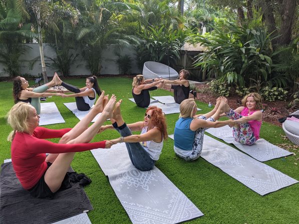 Group of women in an outdoor yoga class practicing partner boat pose on mats on a green lawn in a lush tropical backyard with palm trees