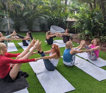 Group of women in an outdoor yoga class practicing partner boat pose on mats on a green lawn in a lush tropical backyard with palm trees